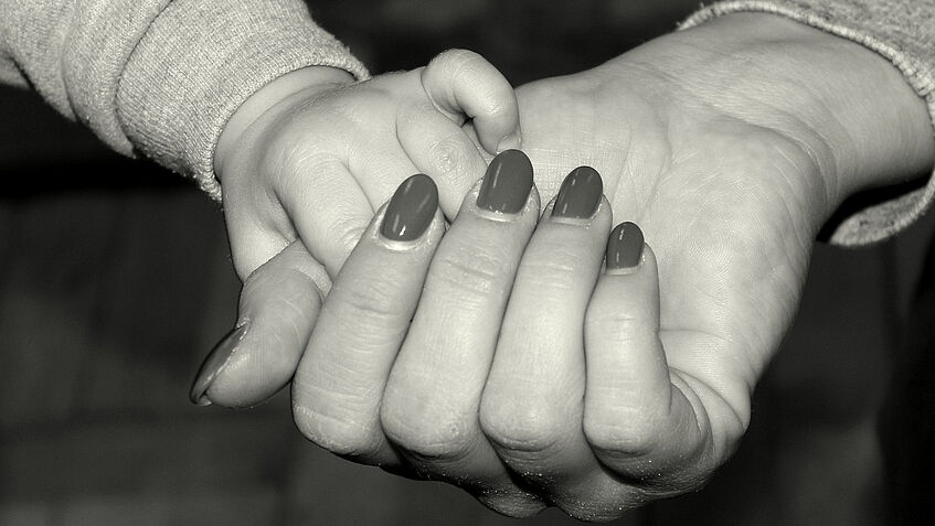 Schwarzweißbild: eine Kinderhand greift in eine halb geschlossene Erwachsenenhand mit lackierten Fingernägeln