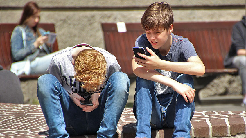 Zwei Buben sitzen auf einer Gehsteigkannte und blicken in ihre Smartphones.