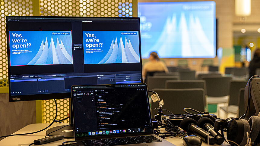 Konferenztitelbild auf Bildschirm, das weiter hinten auf die Leinwand einer Bühne projiziert wird. 