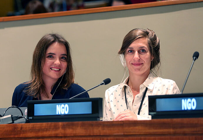 Die beiden Frauen sitzen hinter einem Verhandlungstisch mit dünnen kleinen Mikrofonen. Sie tragen beide einen einseitigen Kopfhörer für die Simultanübersetzung, auf der digitalen Anzeige vor ihnen steht jeweils 'NGO'