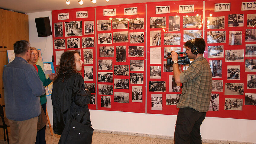 Jürgen Grimm und ein junger Mann mit Kamera die auf eine ältere Frau gerichtet ist, stehen vor einer roten Wand mit vielen kleinen schwarz-weiß Bildern.