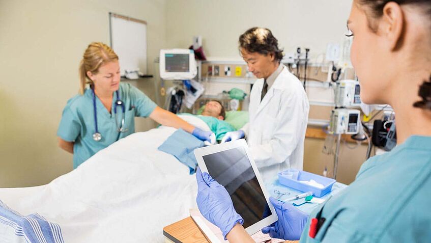 Ein Arzt in weißem Kittel und zwei Pflegerinnen stehen um ein Krankbett. Camera blickt einer Pflegerin über die Schulter, sie hält ein Tablet in der linken Hand.