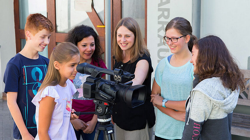 Mehrere Jugendliche stehen vor einer Eingangstür um eine Videokamera auf einem Stativ herum, eine der Jugendlichen blickt durch den Sucher.