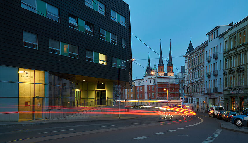 Das Bild zeigt das Multimediazentrum, eine Kirche im Hintergrund und die Straße davor. Durch die langsame Belichtung bei Nacht ziehen die Rücklichter von Autos rote Spuren.
