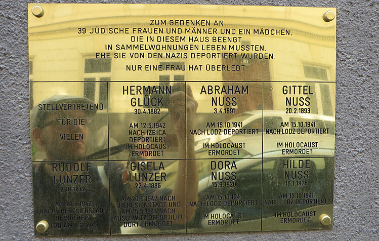 Bild auf eine Messing-Gedenktafel an deportierte JüdInnen eines Hauses, in der Tafel spiegelt sich der Forscher mit Kamera in der Hand