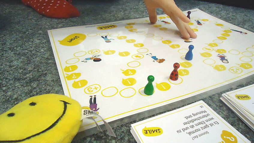 Ein Brettspiel mit drei Hütchen (rot, blau grün) im Vordergrund auf deine weißen Spielbrett mit gelben Feldern. Eine Kinderhand greift ins Spiel, vom nebem den Brett knieenden Kind sieht man nur einen rot besockten Fuß.