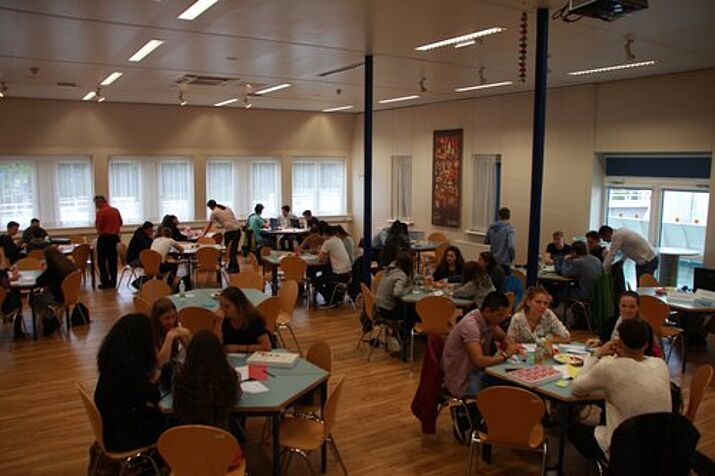 Klassenraum mit Tischen wo SchülerInnen Kleingruppen bilden aus einer Ecke fotografiert.