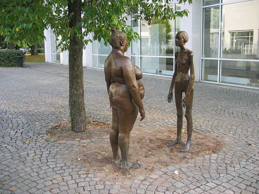 Auf dem gepflasterten Vorplatz, stehen sich eine nakte übergewichtige und eine nakte magersüchtige Frau in Bronze unter einem Baum gegenüber.