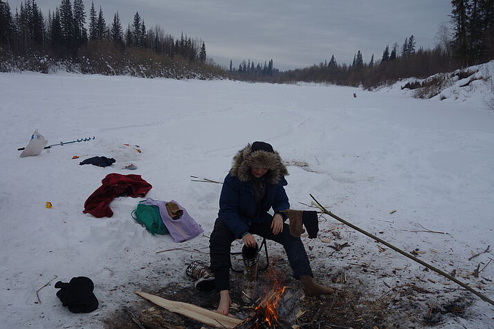 Eine Person sitzt warm angezogen aber Barfuß an einem Feuer, umgeben von Schneelandschaft
