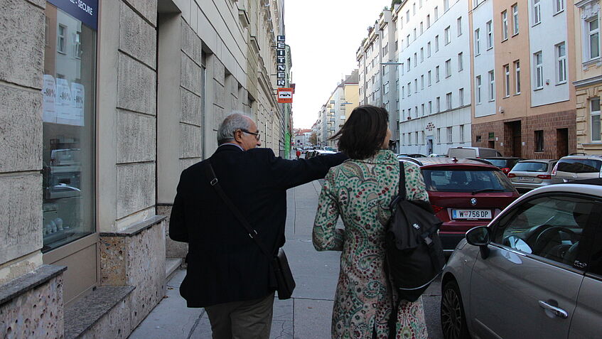 Eine Person und Monika Palmberger sind von hinten abgelichtet während sie am Gehsteig eine Straße entlang gehen. Die Person zeigt mit dem rechten Arm nach rechts über die Straße leicht nach oben, beide blicken in diese Richtung.