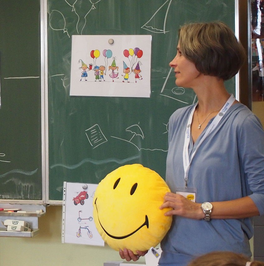 Ulrike Zartler von der Hüfte aufwärts im Portrait. Den Blick nach links gerichtet steht sie vor einer grünen Tafel und hält sie einen Emoji-Smiley in den Händen.